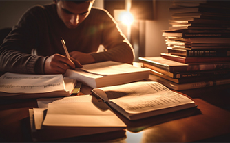 A man sitting at a table with books and a pen, engrossed in studying and writing.