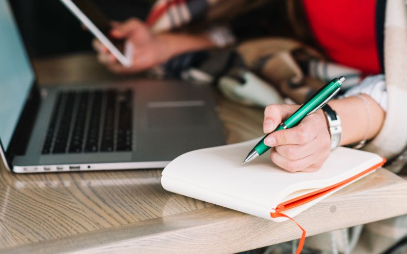 a woman multitasking writing in a notebook with a pen while using a laptop for work or study