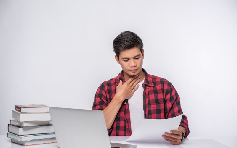 An asian student focused on studying at a desk, surrounded by previous year question papers and using a laptop.