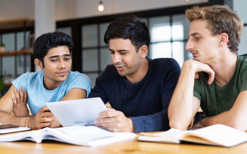 three young men sitting at a table engrossed in a tablet