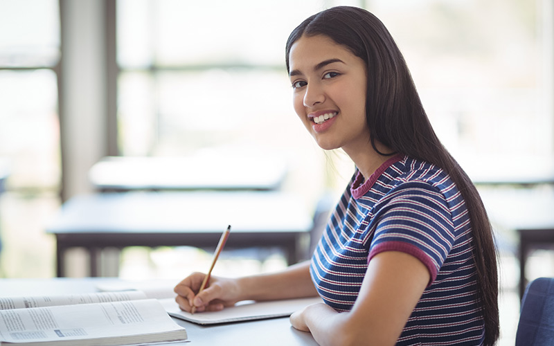 a young woman happily seated at a desk, holding a pen and notebook, ready to jot down her thoughts.