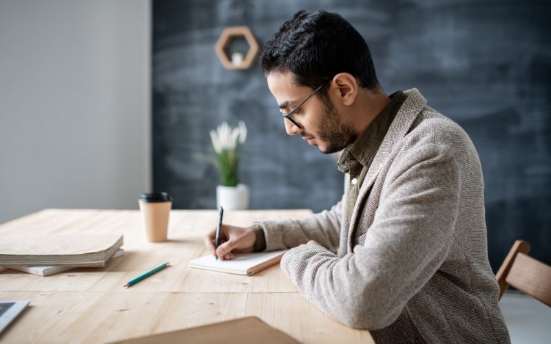 young casual student making notes notebook while sitting by wooden table getting ready with homework